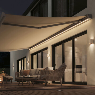 Sonnenschutz- Markisen- Terrassenverschattung mit Markisentuch weiß mit Beleuchtung modernes Haus mit Terasse, Seitenansicht vom Haus mit Terrasse, ausgefahrene Markise über der Terrasse, Markise in weiß, Technik in weiß, Abendansicht, Markise mit Beleuchtung, Hauswand mit Wandlampen beleuchtet